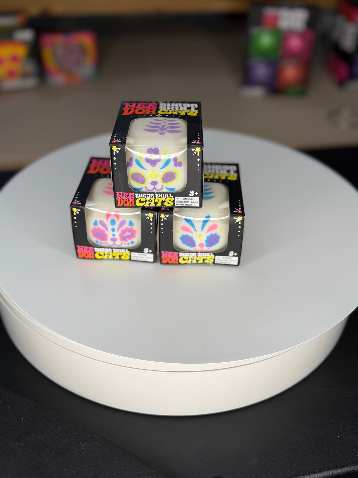 Three boxed cat-shaped items stacked on a round white display platform. The boxes are black with colorful cat illustrations and text including 'MEOW' and 'CATS'.